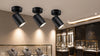 Three black spotlights mounted on a wall, illuminating jewelry displays in a retail store setting.