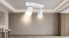 Modern minimalist room with a white dual ceiling spotlight. Soft lighting highlights neutral walls, a textured rug, and a small table with vases, creating a calm atmosphere.