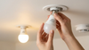 Hands are shown installing a white LED light bulb into a ceiling socket. Another bulb nearby is lit, casting a warm glow. The scene conveys careful precision.