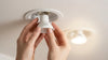 Hands are installing an LED light bulb into a recessed ceiling fixture. The bulb is white with a gray logo, and another bulb in the background is lit.
