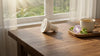 A white light bulb lies on a sunlit wooden table next to a tray with a coffee cup and morning glory flower. A window reveals a serene garden view.