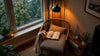 A cozy chair with a book and a lamp beside it, positioned in front of a bright window.